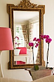 Reflection of pale, country-house living room in mirror with antique gilt frame; pink lampshade and orchids in foreground: feminine atmosphere
