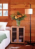 Kleiner Schrank mit Drahtgittertüren als Nachttisch neben Doppelbett; darauf ein prachtvoller Blumenstrauss mit orangen Blüten