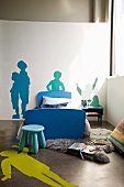 Zimmerecke mit blauem Hocker vor Einzelbett mit blauem Bettgestell und Schablonenmalerei mit Menschenmotiven auf Boden und Wand
