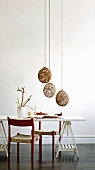 Red-painted kitchen chair at white table on wooden trestles and pendant lamps with spherical, basketwork shades made from natural materials