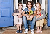 Zwei junge Frauen und ein junger Mann sitzen mit Gemüse vor der Tür