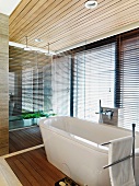 Free standing bathtub in front of a bank of windows with closed blinds in a modern bathroom with a wooden ceiling