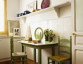 Simple kitchen table and green-painted chairs against tiled wall and below white shelf