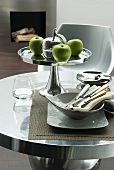 Green apples on silver fruit stand and bowl of cutlery on round table