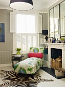 Patterned chaise longue below window with white, interior shutters and zebra-skin rug on white wooden floor in front of fireplace in traditional interior