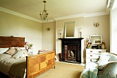 Traditionally furnished bedroom with inlaid wooden bed, white chest of drawers with mirror next to old open fireplace with blue Baroque sofa below window