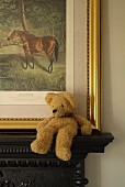 Beige teddy bear sitting slumped on dark mantelpiece in front of gilt-framed picture of horse in countryside leaning on wall