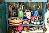 Blue wooden cupboard with paintbrushes, pens and sewing materials in patterned boxes and jugs