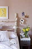 Bed with white bed linen and DIY headboard of birch branches with nesting box on wall. DIY grey bedside table with white, wooden breakfast tray.