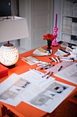 Orange work desk with pens and markers; at the edge a lighted table lamp, roses and a pink candle stick