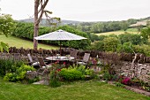 Idyllic seating area on corner terrace against stone garden wall with table, chairs and parasol and view across rolling English landscape in background
