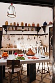 Einblick in schlichte orientalische Küche mit massivem Küchentisch aus Stein und hohen Holzhockern, dahinter massiver Spültisch, Wandleiste mit aufgehängten Küchenutensilien und Konsolenregale mit gefüllten Einmachgläsern