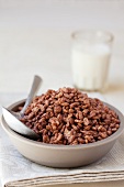 A bowl of chocolate rice puffs with a glass of milk in the background