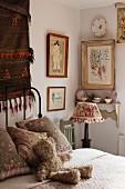 Corner of feminine bedroom with framed pictures and patterned china on wall bracket behind nostalgic bedside lamp on wooden chair; cuddly teddy bear waiting on cosy bed