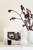 Romantischer weißer Bilderrahmen mit einer schwarz-weiß Fotographie von Hund mit Dame auf einem weiß lackierten Schrankelement mit schlichten weißen Milchglasblumenvasen daneben; ein rötlicher Zweig schenkt einen Farbakzent