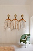 Antique Hungarian linen smocks and shirts hung decoratively on a white wall above a green Lloyd Loom chair