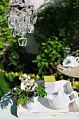 A chandelier above a tray with a porcelain teapot, tea caddy and a jug of flowers in a sunny garden