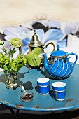 A blue porcelain teapot with silver decoration and matching tea tumblers on an old, round, metal garden table