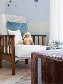 Armchair with cushions against wall painted pale blue and white and large, cubic pouffe with brown corduroy cover in corner of nursery with various soft toys decorating the pleasant room