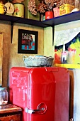 Roter Retrokühlschrank unter kleinen Gemälden und einfachem Holzbord mit bunten Dosen in einer Küchenecke