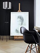 Antike Staffelei mit darauf stehendem, modernen Portrait vor schwarzer Wand; im Vordergrund schwarze Charles Eames Stühle