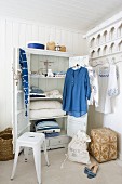 Offener Kleiderschrank mit an der Tür hängenden Kleidungsstücken in Blau und Weiß; rundherum Körbe, ein Wäschesack und ein weisser Metallhocker