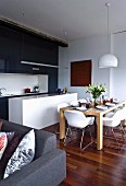 Open-plan interior with set dining table in front of black and white fitted kitchen; corner of grey sofa in foreground