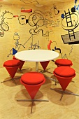 Modern, plywood paneled conference room with a white, round table and stools upholstered in orange fabric in front of a trendy wall decorated with cartoons (Red Bull Zentrale, Amsterdam)