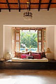 Floor cushions and scatter cushions on wooden platform below open window with shutters and view of pool in garden