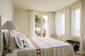 Bedside lamp with white lampshade next to double bed in country-style bedroom with white, floor-length curtains at windows