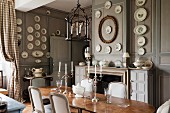Candlesticks on antique table and pale, upholstered chairs in grand dining room with wall plates on grey-painted, wood-panelled walls