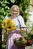 Frau mit Sonnenblumen und Gemüsekorb vor dem Bauernhaus