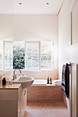 Base unit with protruding washbasin and fitted bathtub below windows with louver blinds in narrow bathroom