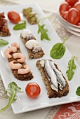 Slices of bread with sardines, shrimps and squid on a rectangular platter with lettuce leaves and cherry tomatoes