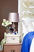 Bedside table on stacked trunks next to inflatable bed against patterned wallpaper