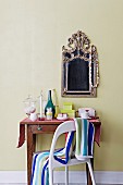 Striped towel on back of chair in front of antique console table below mirror with ornate metal frame on pastel green wall
