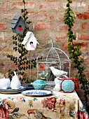 Stylised fruit on plate and china bird in front of birdcage on table against brick wall