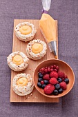 A board with four meringue nests filled with orange cream, next to a piping bag of orange cream and a bowl of fresh berries