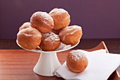 Several doughnuts coated with sugar glaze or icing sugar, on a cake stand against a purple background