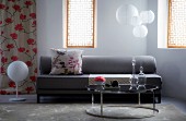 Graues Sofa mit geblümtem Zierkissen und dahinter hängenden Papierlampen; davor ein glänzender Couchtisch mit Glaskaraffen