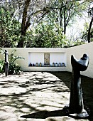 Modern stone sculptures in courtyard and vessels in niche of garden wall in front of jungle-like woodland