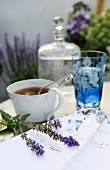 Tasse mit Tee-Ei-Zange und Minzblüte; blaues Kristallglas und Wasserkarraffe unscharf im Hintergrund