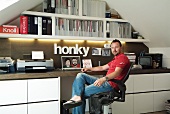 Man sitting at workstation in attic room with fitted shelving and white cupboard elements below worksurface