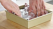 A baking tin being lined with aluminium foil