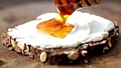 A slice of wholemeal bread topped with cream cheese being drizzled with honey