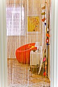 Blick durch luftigen Fransenvorhang auf farbenfrohe Zimmerecke mit orangefarbenem Sessel
