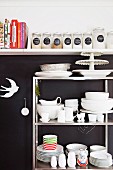 Metal shelves of white crockery against black wall below white, wall-mounted kitchen shelf of cookery books and storage jars