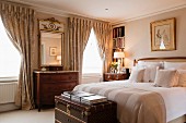 Antique chests of drawers, draped curtains and elegant trunk at foot of double bed in traditional bedroom