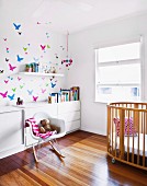 Classic rocking chair next to cot and white sideboard against wall with butterfly motifs in sunny nursery