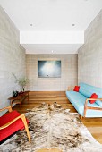 Animal-skin rug on wooden floor between armchair with red cover and blue, 50s-style sofa in anteroom with pale grey stone walls
